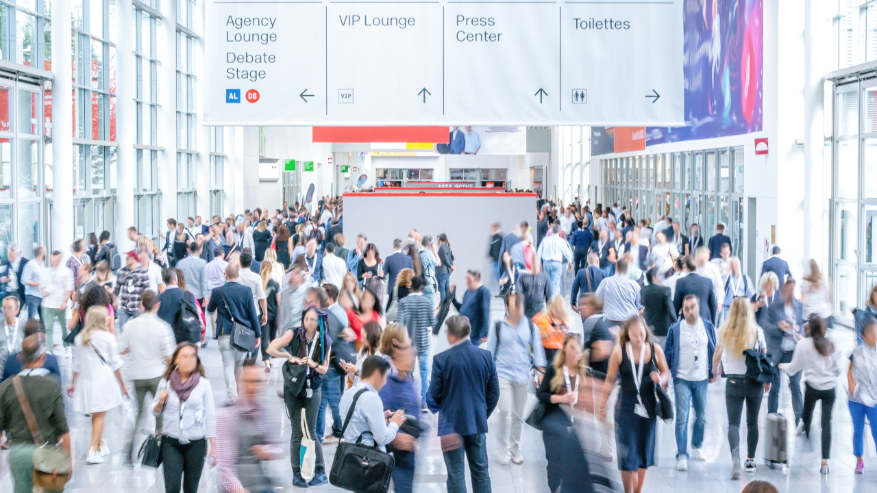 Crowded convention center hallway with people walking. Overhead signs indicate lounge areas and facilities. Bright, bustling atmosphere.