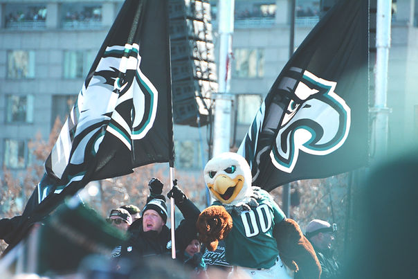 Eagle mascot and fans wave large black flags with logos at an outdoor rally. The city building backdrop adds a festive atmosphere.